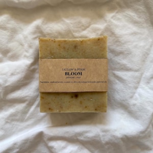 May include: A rectangular bar of soap with a speckled beige texture, wrapped in a tan paper band. The label reads TALLOW & FORM, BLOOM, jasmine rose, and lists ingredients including rendered tallow, coconut oil, olive oil, sodium hydroxide, and essential oils.