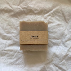 Op de afbeelding: Een rechthoekig stuk beige zeep verpakt in een bruin papieren label. Het label bevat de tekst TALLOW & FORM, EMBER, clove, sandalwood en een ingrediëntenlijst. De zeep heeft een glad, solide uiterlijk met een licht getextureerde rand.