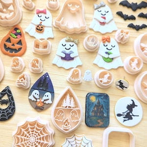 Könnte beinhalten: Eine Sammlung von Halloween-Ausstechformen aus Polymer-Ton und fertigen Anhängern, darunter Geister-, Kürbis-, Hexen-, Spinnennetz- und Spukhaus-Motive in verschiedenen Größen, Farben und Designs, angeordnet auf einer hellen Holzoberfläche.