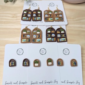 May include: Handmade polymer clay earrings and stud earrings in the shape of bookshelves. The earrings are brown with miniature books, plants, and other decorative items. The stud earrings feature a small bookshelf, a plant, and a stack of books. The text "Small and Simple Joy" is printed on the cards.