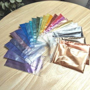 May include: A collection of 20 small bags of iridescent mica powder in various colors, including gold, silver, blue, green, purple, and red. The bags are arranged in a fan shape on a wooden surface.