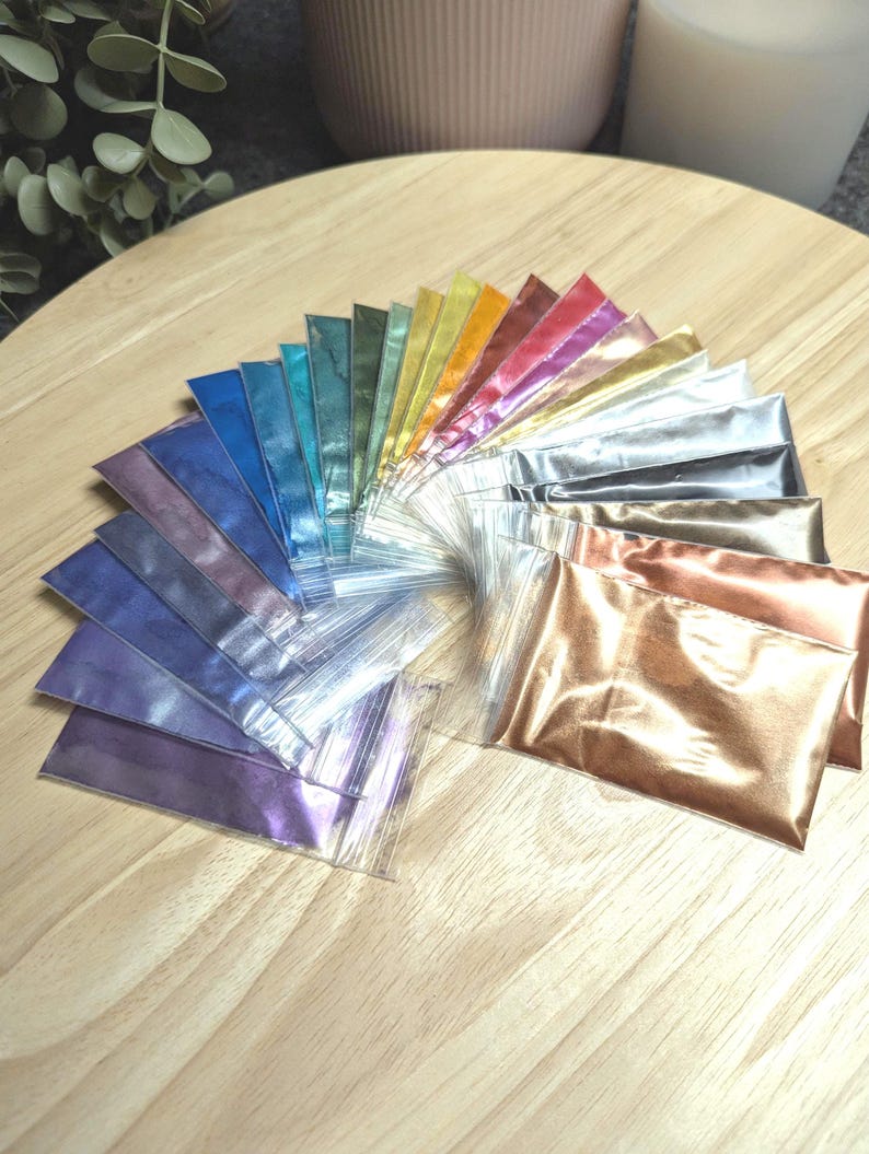 May include: A collection of 20 small bags of iridescent mica powder in various colors, including gold, silver, blue, green, purple, and red. The bags are arranged in a fan shape on a wooden surface.