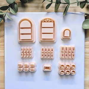 Puede incluir: Una colecci&oacute;n de cortadores de galletas de color melocot&oacute;n claro en varias formas, incluyendo ventanas arqueadas, formas rectangulares y dise&ntilde;os redondeados. Los cortadores est&aacute;n dispuestos sobre una superficie blanca con follaje verde en el fondo.