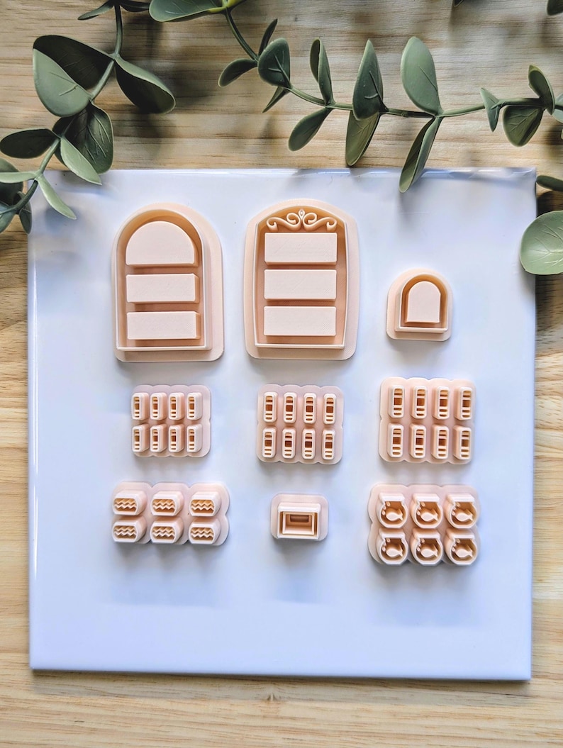 Puede incluir: Una colecci&oacute;n de cortadores de galletas de color melocot&oacute;n claro en varias formas, incluyendo ventanas arqueadas, formas rectangulares y dise&ntilde;os redondeados. Los cortadores est&aacute;n dispuestos sobre una superficie blanca con follaje verde en el fondo.