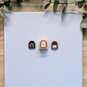 Puede incluir: Tres dijes de arcilla polim&eacute;rica en miniatura con forma de arcos. Un arco presenta una pila de libros, otro es de color neutro y el tercero tiene una peque&ntilde;a planta en maceta. Los dijes est&aacute;n sobre una superficie blanca.