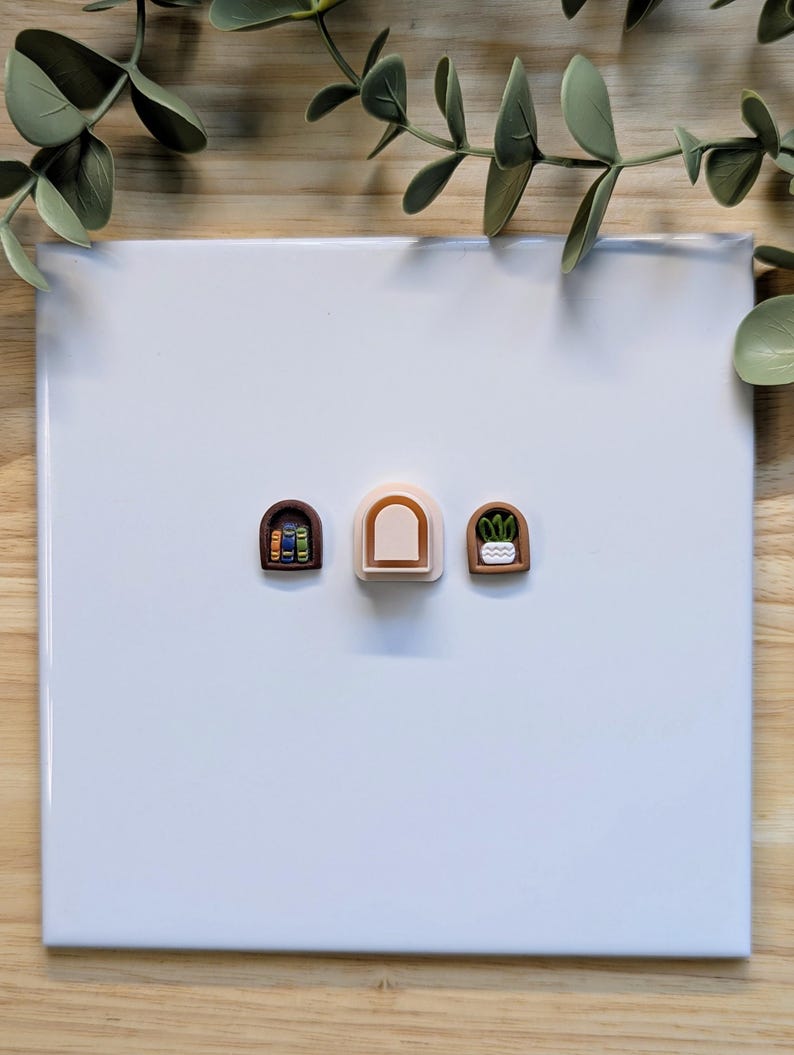 Puede incluir: Tres dijes de arcilla polim&eacute;rica en miniatura con forma de arcos. Un arco presenta una pila de libros, otro es de color neutro y el tercero tiene una peque&ntilde;a planta en maceta. Los dijes est&aacute;n sobre una superficie blanca.
