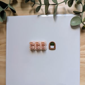 Puede incluir: Un conjunto de cortadores de arcilla polim&eacute;rica en color melocot&oacute;n claro. Los cortadores incluyen tres juegos de l&iacute;neas onduladas y una peque&ntilde;a ventana arqueada con un dise&ntilde;o de planta en maceta. Dispuestos sobre una superficie blanca.