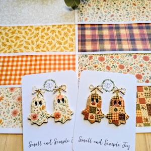 May include: Two pairs of ghost-shaped earrings on white display cards labeled Small and Simple Joy. One pair features a floral autumn pattern, while the other displays a patchwork quilt design. Each ghost has black oval eyes and a gold bow topper.