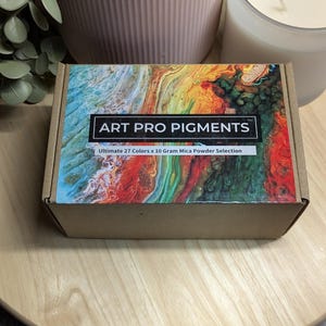 May include: A cardboard box with a colorful abstract design on the top. The box is labeled "ART PRO PIGMENTS" and "Ultimate 27 Colors x 10 Gram Mica Powder Selection".