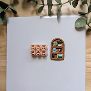 Puede incluir: Colecci&oacute;n de imanes en miniatura hechos a mano. Un juego presenta seis peque&ntilde;os frascos transparentes con l&iacute;quido naranja. El otro es una estanter&iacute;a marr&oacute;n con libros, una planta y una imagen enmarcada. Los imanes est&aacute;n sobre una superficie blanca.