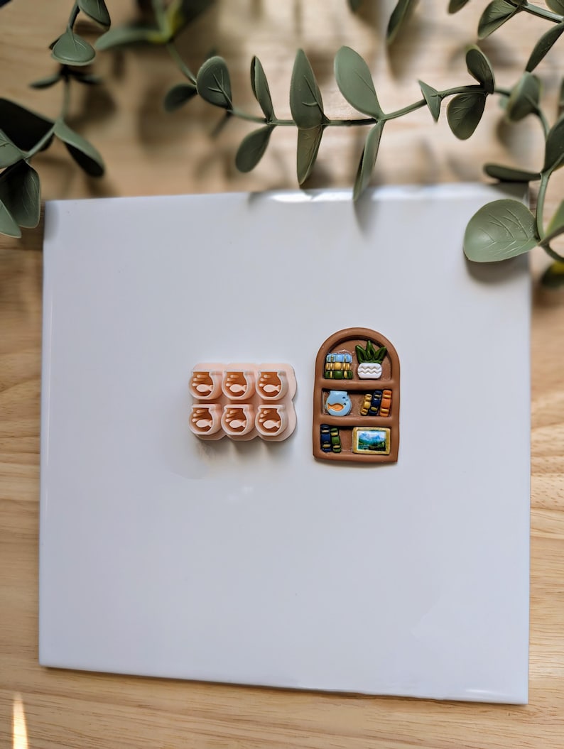Puede incluir: Colecci&oacute;n de imanes en miniatura hechos a mano. Un juego presenta seis peque&ntilde;os frascos transparentes con l&iacute;quido naranja. El otro es una estanter&iacute;a marr&oacute;n con libros, una planta y una imagen enmarcada. Los imanes est&aacute;n sobre una superficie blanca.