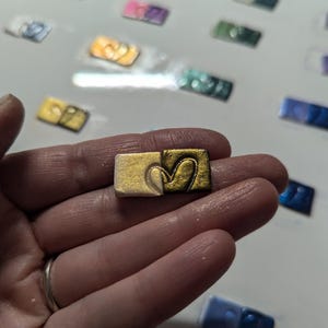 May include: A small, square, gold-colored paint pan with a heart-shaped design. The pan is half-filled with gold paint and half-filled with white paint.
