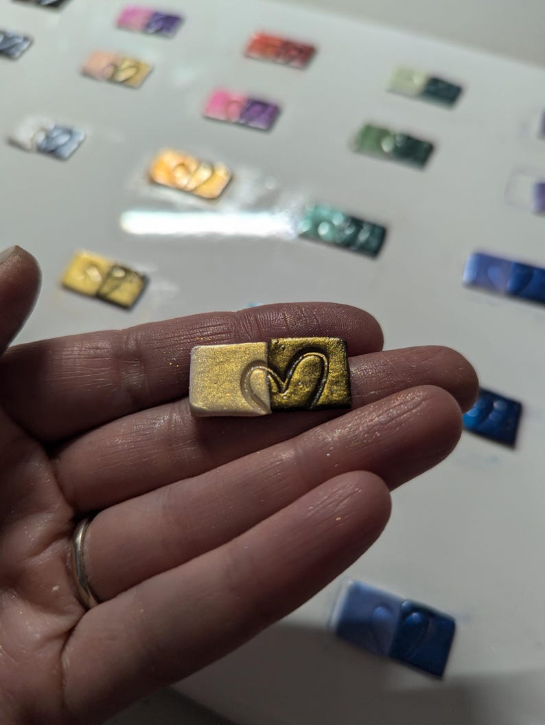 May include: A small, square, gold-colored paint pan with a heart-shaped design. The pan is half-filled with gold paint and half-filled with white paint.