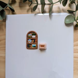 Puede incluir: Un im&aacute;n en miniatura con forma de estanter&iacute;a marr&oacute;n con libros peque&ntilde;os, una planta y obras de arte. Junto a &eacute;l hay un peque&ntilde;o im&aacute;n rectangular de color marr&oacute;n claro. Los imanes est&aacute;n sobre una superficie blanca, con ramitas verdes con hojas en el fondo.