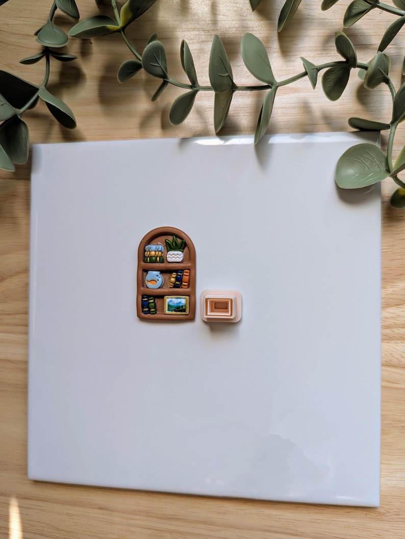 Puede incluir: Un im&aacute;n en miniatura con forma de estanter&iacute;a marr&oacute;n con libros peque&ntilde;os, una planta y obras de arte. Junto a &eacute;l hay un peque&ntilde;o im&aacute;n rectangular de color marr&oacute;n claro. Los imanes est&aacute;n sobre una superficie blanca, con ramitas verdes con hojas en el fondo.