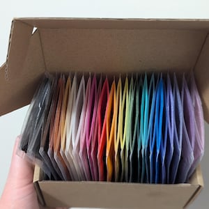 May include: A cardboard box filled with small, individually packaged bags of colorful glitter. The glitter is arranged in a rainbow pattern.