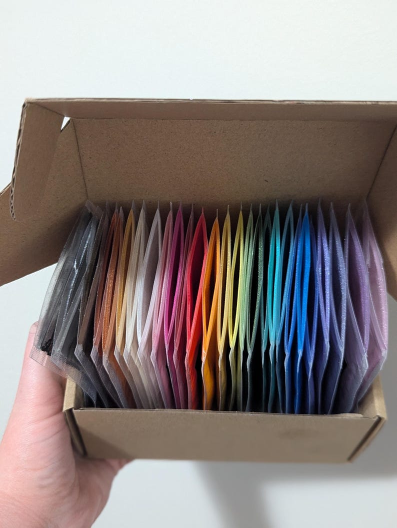 May include: A cardboard box filled with small, individually packaged bags of colorful glitter. The glitter is arranged in a rainbow pattern.