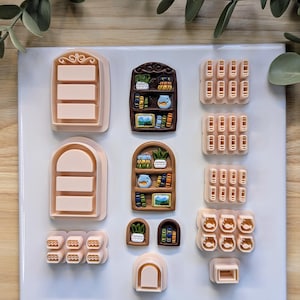 May include: A collection of light pink polymer clay cutters in various shapes, including bookshelves, windows, and stacks of books. The cutters are arranged on a white surface, with green foliage in the background.