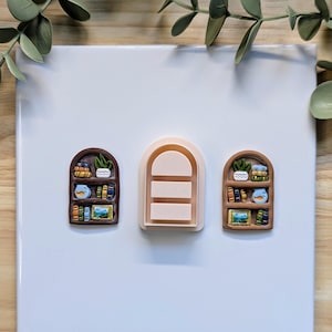 Puede incluir: Tres cortadores de arcilla polim&eacute;rica con forma de estanter&iacute;a. Dos est&aacute;n detallados con libros en miniatura, plantas y obras de arte. El tercero es una forma de arco simple con tres l&iacute;neas horizontales. Los cortadores son marrones, beige y de varios colores.