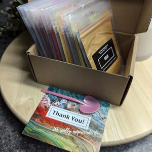 May include: A cardboard box filled with small plastic bags containing various colors of mica powder. The box is open and the bags are arranged in rows. The top bag is labeled "Gold". A thank you card with a colorful abstract design is on a wooden surface.
