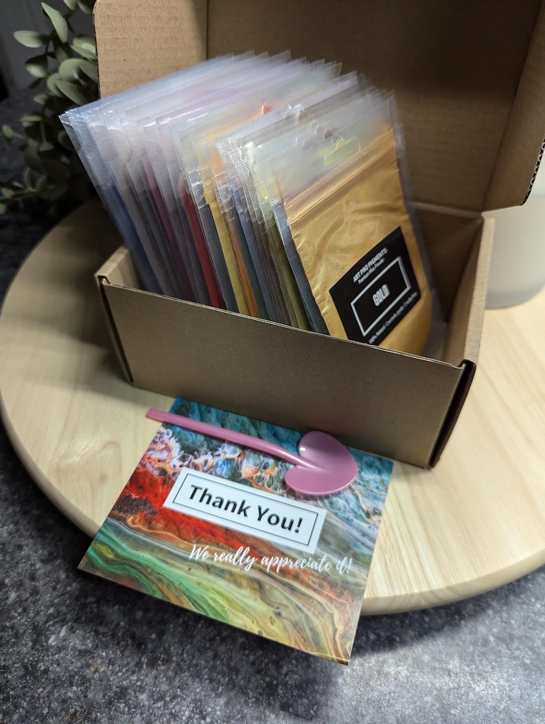 May include: A cardboard box filled with small plastic bags containing various colors of mica powder. The box is open and the bags are arranged in rows. The top bag is labeled "Gold". A thank you card with a colorful abstract design is on a wooden surface.