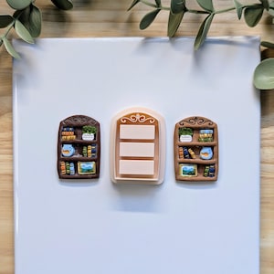 Puede incluir: Tres objetos decorativos con forma de estanter&iacute;a. Dos son marrones con libros, plantas y un p&aacute;jaro. El objeto central es un rect&aacute;ngulo rosa claro con tres l&iacute;neas horizontales. Los objetos est&aacute;n sobre una superficie blanca con hojas verdes en el fondo.