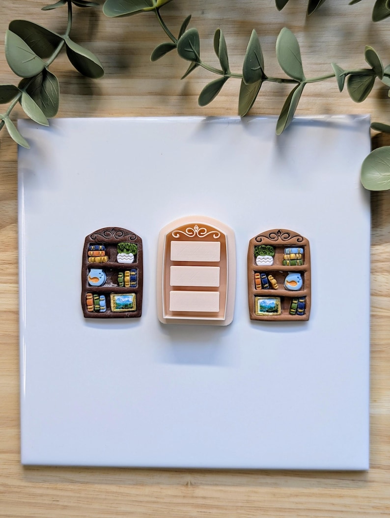 Puede incluir: Tres objetos decorativos con forma de estanter&iacute;a. Dos son marrones con libros, plantas y un p&aacute;jaro. El objeto central es un rect&aacute;ngulo rosa claro con tres l&iacute;neas horizontales. Los objetos est&aacute;n sobre una superficie blanca con hojas verdes en el fondo.