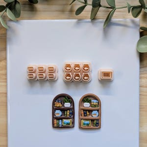 Puede incluir: Una colecci&oacute;n de creaciones de arcilla polim&eacute;rica en miniatura, que incluyen dise&ntilde;os en forma de hamburguesa, pecera y rectangulares. Tambi&eacute;n hay dos piezas en forma de estanter&iacute;a arqueada, cada una con libros en miniatura detallados, plantas y una pecera. Los art&iacute;culos est&aacute;n dispuestos sobre una superficie blanca.