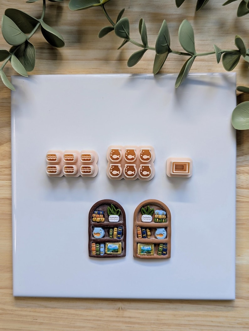 Puede incluir: Una colecci&oacute;n de creaciones de arcilla polim&eacute;rica en miniatura, que incluyen dise&ntilde;os en forma de hamburguesa, pecera y rectangulares. Tambi&eacute;n hay dos piezas en forma de estanter&iacute;a arqueada, cada una con libros en miniatura detallados, plantas y una pecera. Los art&iacute;culos est&aacute;n dispuestos sobre una superficie blanca.