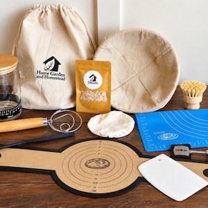 May include: A sourdough bread baking kit including a glass jar with a bamboo lid, a beige canvas drawstring bag, a round proofing basket, a wooden-handled dough whisk, a blue silicone mat, a scoring lame, a white plastic scraper, and a tan mat.