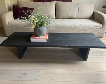 May include: A black wooden coffee table with a simple design and a book on top. A vase with white flowers sits on the book. The table is in front of a light-coloured sofa.