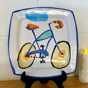 May include: Square ceramic plate with a whimsical bicycle design. The plate features a white background with a blue border and a colorful bicycle illustration in shades of blue, orange, and yellow. The plate is displayed on a black stand.