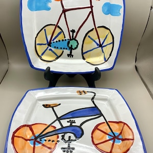 May include: Two square ceramic plates with hand-painted bicycle illustrations. The top plate features a red bicycle with yellow wheels and blue clouds. The bottom plate displays a blue bicycle with orange wheels. Both plates have blue rims.