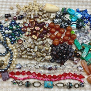 May include: A large collection of assorted loose beads and jewelry components in various shapes, sizes, and colors, including strands of pearls, faceted red and pink beads, smooth orange stones, green rectangular pieces, and patterned glass beads.