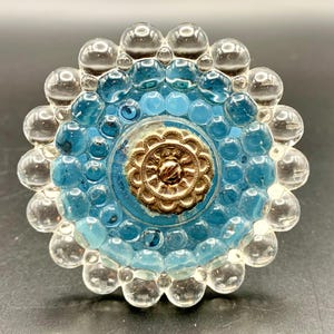 May include: A decorative glass knob with a floral design. The knob features a central gold-colored medallion surrounded by blue and clear glass bubbles, creating a flower-like shape. The knob is likely for furniture or a cabinet.