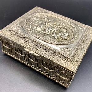 May include: Silver-toned rectangular box with ornate relief detailing. The lid features an oval scene with figures, surrounded by a rope-like border. The sides have a repeating garland design, creating a vintage aesthetic.