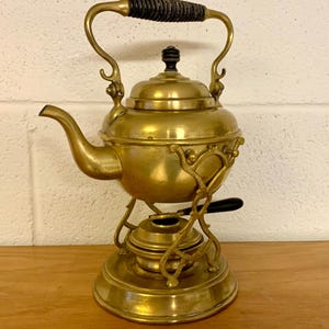 May include: Antique brass tea kettle with a black handle and a decorative base. The kettle has a curved spout and a lid with a small black knob. The base has an ornate design. The kettle is sitting on a wooden surface.