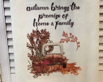 May include: A white kitchen towel with brown stripes features a vintage truck with fall leaves and the text "autumn brings the promise of home & family".