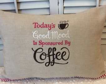 May include: A beige linen pillow with red and white striped trim. The pillow has embroidered text that reads "Today's Good Mood Is Sponsored By Coffee" with a stylized coffee cup and steam.