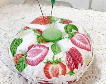 May include: A round strawberry-themed pin cushion with a white lace trim. The cushion features a white fabric with a pattern of red strawberries and green leaves. Two pins with pink and green heads are inserted into the cushion.