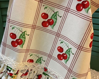 May include: A cream-colored apron with a cherry pattern in a grid design. The apron has a ruffled bottom with a vintage kitchen utensil print and lace trim. The cherries are red with green leaves.
