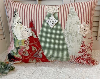 May include: Rectangular decorative pillow with a red and white striped background. The pillow features three appliqued Christmas tree shapes in red floral, green gingham, and toile fabrics, adorned with white lace snowflakes.