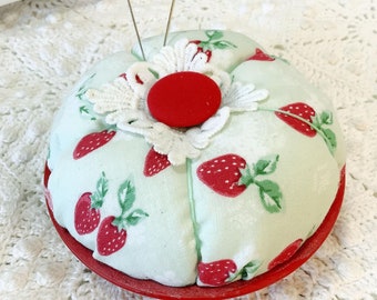 May include: A green and white pincushion shaped like a strawberry with red strawberries printed on it. The pincushion has a red center and a white lace trim. Two pins are stuck in the top of the pincushion.