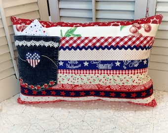 May include: A rectangular decorative pillow with a patriotic theme. The pillow features a denim pocket with a heart-shaped patch and lace trim. The pillow has red, white, and blue fabric stripes with star and cherry patterns.