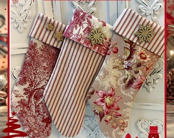 May include: Three Christmas stockings featuring a red and white toile pattern, a red and white striped pattern, and a floral print. Each stocking has a folded cuff adorned with gold glitter snowflake ornaments. The text MERRY CHRISTMAS is visible.
