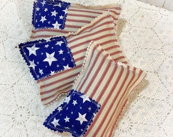 May include: Four patriotic-themed decorative pillows. Each pillow features a blue square with white stars, reminiscent of the American flag, and red and white striped fabric. The pillows have a rustic, homespun aesthetic.