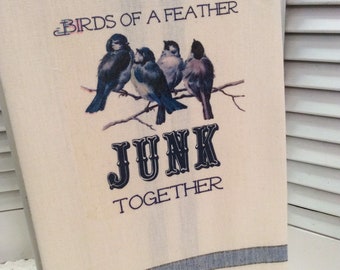 May include: A white tea towel with blue stripes at the bottom. The towel has a printed image of four blue birds on a branch with the text "Birds of a feather junk together".