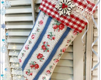 May include: A Christmas stocking featuring a red and white gingham cuff adorned with a white lace trim and a wooden snowflake embellishment. The body of the stocking displays a pattern of blue vertical stripes and small red floral clusters.