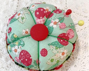 May include: A round fabric pincushion featuring a light mint green background with a pattern of red strawberries and white flowers. A solid red fabric-covered button sits in the center, and two sewing pins with red and yellow heads are inserted.
