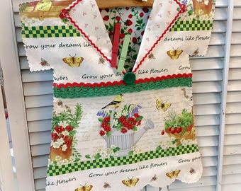 May include: A fabric wall hanging shaped like a shirt, featuring a cream background with floral, butterfly, and watering can prints. It includes the text "Grow your dreams like flowers," a green crochet trim, a green button, and two clothespins.